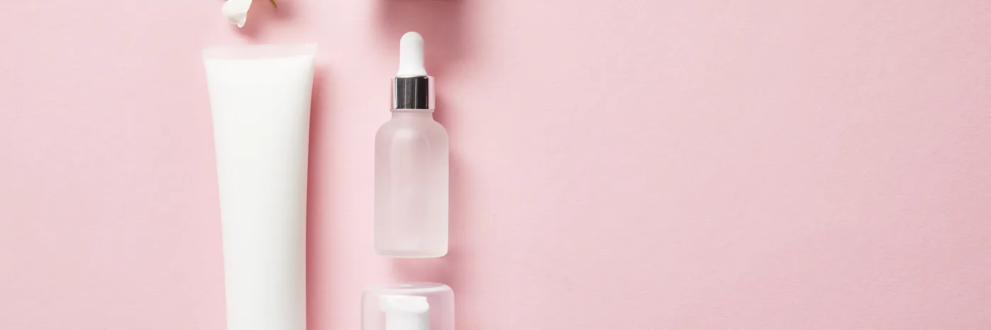 An overhead flat lay of unbranded white cosmetic containers on a soft pink background: a cream tube, a dropper bottle, a pump bottle and two lidded jars beside a jasmine flower.